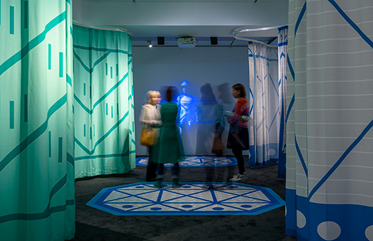 People walking between colourful curtains in the Radical Rooms exhibition space